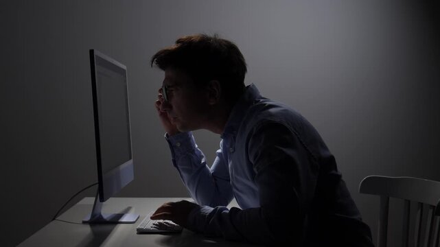 Focused man in glasses working at a computer in a dimly lit office, experiencing eye strain and fatigue from prolonged screen time and overwork
