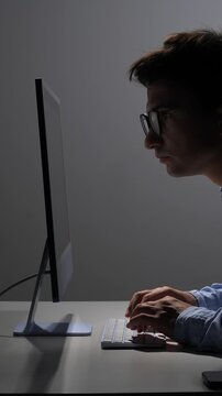 Vertical video man wearing glasses intently focusing on his computer screen while typing on a keyboard, working or programming late at night in a dark office setting