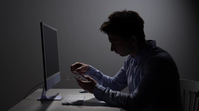 Man feeling stressed and frustrated, holding tangled in ear headphones while sitting at a desk and using a computer in a dimly lit environment, symbolizing technology induced challenges
