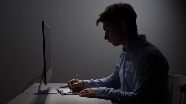 Man concentrating intensely on work at a desk in a dark office, writing on a clipboard while a monitor glows in the background, symbolizing dedication and demanding tasks