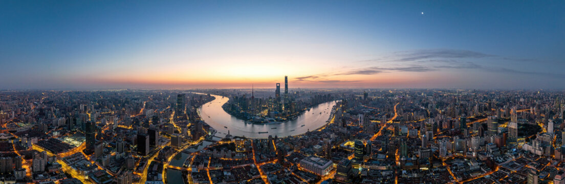 A wide panoramic aerial view of a sprawling urban city with a winding river, illuminated buildings and roads, at dawn with a clear sky.