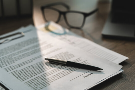 Close-up of a pen on printed agreement papers beside a signature line, clipboard and eyeglasses, showing contract approval preparation and formal business office documentation workflow.