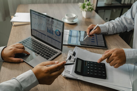 Business colleagues comparing financial data on laptop and tablet dashboards with a calculator and printed reports, showing analysis and verification workflow at an office desk.