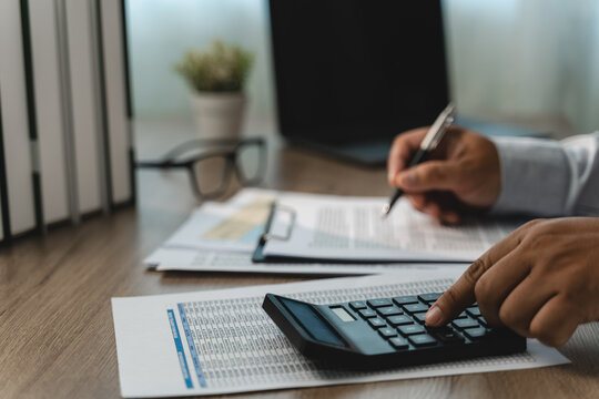 Hands using a calculator and pen with printed financial tables at an office desk, showing expense calculation, detailed budget review and a financial document analysis workflow for business.