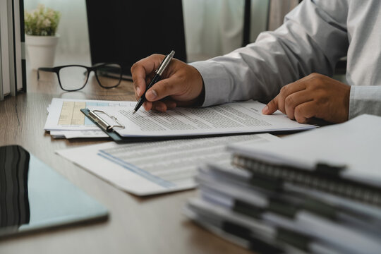 One hand holds a pen while another points to terms on printed agreement documents, showing contract terms review and business document approval workflow at an office desk.