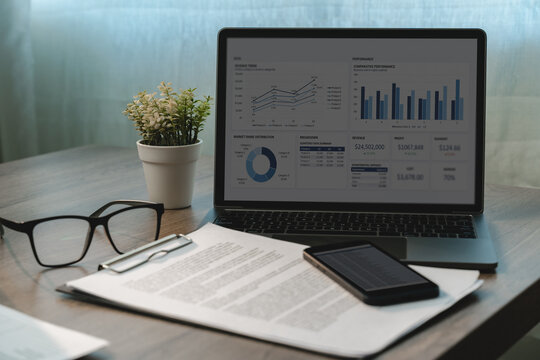 Laptop displaying business performance charts beside printed reports, smartphone and eyeglasses on an office desk, showing financial data analysis, revenue review and reporting workflow.