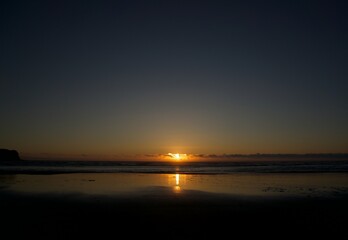 Fototapeta premium Wide Angle Landscape of Oregon Coast at Sunset with Sun Dipping Below Low Cloud Horizon and Perfect Light Beam Reflection on Wet Flat Sandy Beach