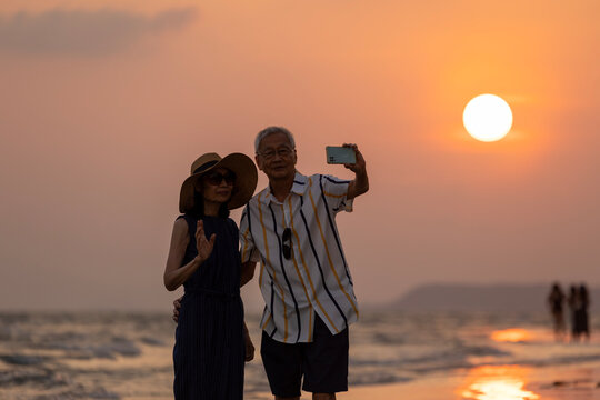 Silhouette  of Asian senior couple taking selfie while watching at the sunset sea beach in summer for healthy relationship and happy loving marriage life in vacation retirement years