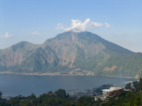 Aerial view of Mount Abang rising above the serene waters of Lake Batur, surrounded by lush green slopes and distant village structures in Kintamani, Bali, Indonesia.