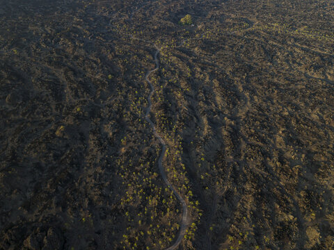 Aerial view of the volcanic landscape of Mount Batur with a winding dirt path cutting through dark solidified lava fields and sparse green vegetation in Kintamani, Bali, Indonesia.