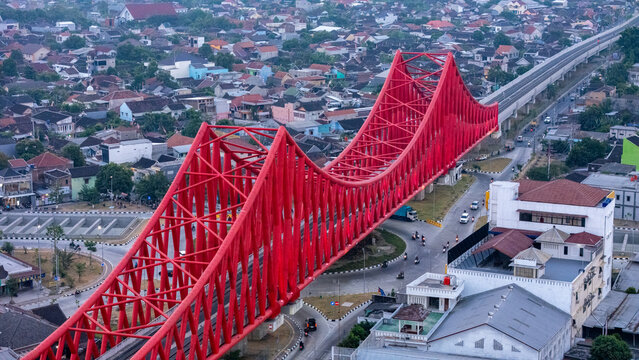 Aerial view of the vibrant red Tirtonadi Bridge spanning over a busy road intersection with surrounding urban rooftops and city infrastructure in Surakarta, Central Java, Indonesia.
