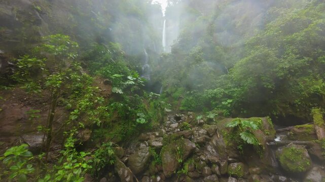 FPV drone flight through misty rainforest canyon in Costa Rica revealing tall waterfall falling into turquoise river between mossy cliffs lush tropical foliage and rocky mountain stream.
