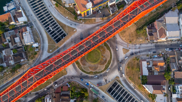 Aerial view of a vibrant red steel truss bridge spanning over a busy roundabout with urban traffic and surrounding buildings in Surakarta, Central Java, Indonesia.