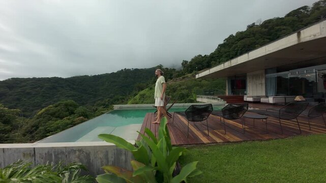 FPV drone flight around modern concrete villa in Costa Rica where father and son walk barefoot along wooden deck beside infinity pool with rainforest hills and ocean horizon behind.