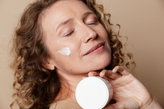 Smiling middle aged woman applies white cream to her cheek. Close up on a neutral background. Skincare, wellness, and selfcare concept with natural, unretouched skin.