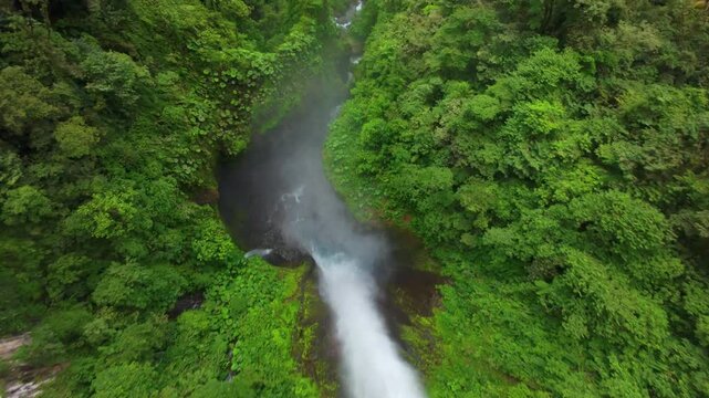 FPV drone dives through misty jungle canyon in Costa Rica revealing a waterfall plunging into a rocky river surrounded by dense tropical rainforest foliage and vibrant green treetops.