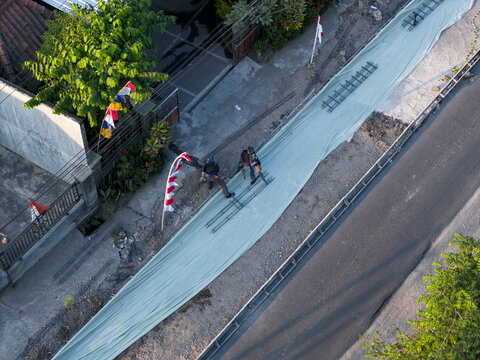 Aerial view of concrete road construction with workers placing steel reinforcement bars on a protective sheet along the roadside in Kecamatan Manisrenggo, Jawa Tengah, Indonesia.