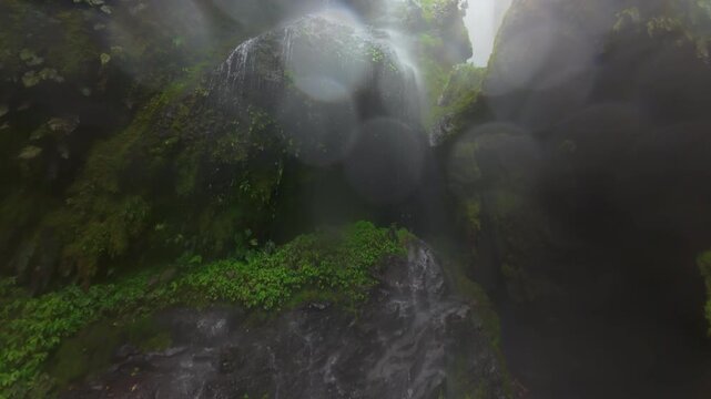 FPV drone flight through misty gorge in Costa Rica rainforest passing mossy rock walls dripping water and diving beside tall waterfall plunging into lush tropical jungle canyon below.