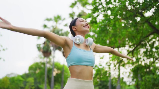 Asian woman wearing wireless headphones and activewear, standing in a green public park enjoying fresh air with a relaxed and refreshed expression. Outdoor fitness and wellness lifestyle concept