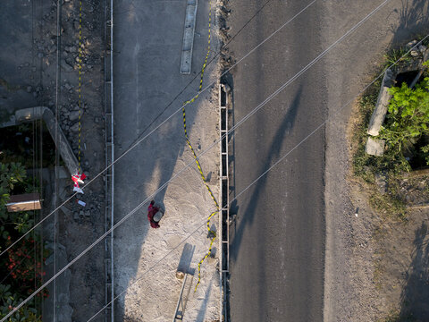 Aerial view of concrete road construction with a worker in red clothing, yellow caution tape, and power lines casting long shadows in Kecamatan Manisrenggo, Jawa Tengah, Indonesia.
