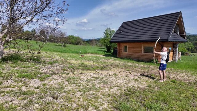 Outdoor archery session featuring a man drawing, releasing and preparing the next arrow. Beautiful countryside, wooden cabin, blue sky and active lifestyle.