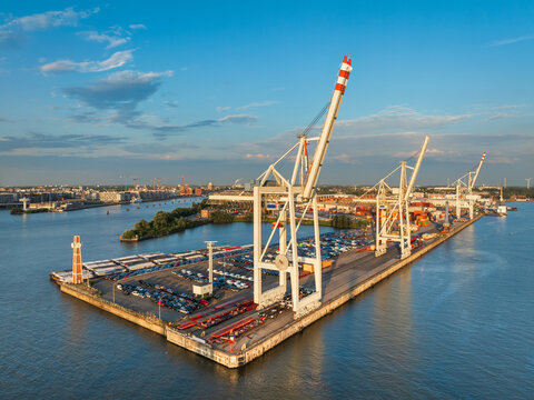 Aerial view of the Port of Hamburg with large container cranes, rows of parked cars on the terminal, and the river Elbe under a clear blue sky in Hamburg, Hamburg, Germany.