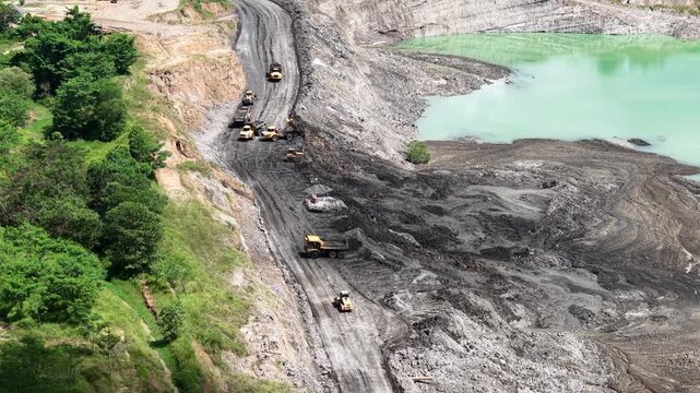 An aerial view of a former coal mine pit filled with blue water shows haul trucks operating on a road at the lake's edge, with plumes of smoke rising nearby
