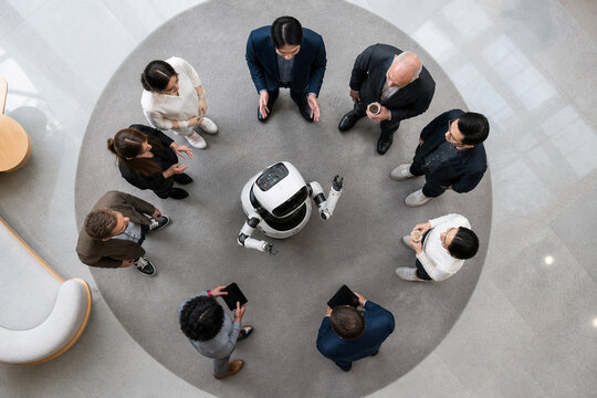 Top view of a small robot surrounded by a diverse team of professionals in business attire, gathered around a circular rug in a bright modern atrium with glass floors for a collaborative discussion.