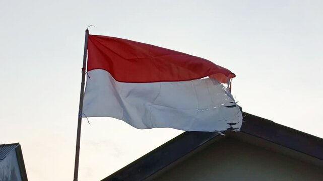 Video footage of a torn Indonesian flag waving on a pole above a house rooftop against pale sky.