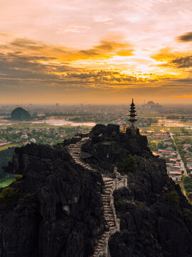 Aerial view of Mua Cave with its stone staircase winding up the rocky peak, a pagoda at the summit, and a person standing nearby under a vibrant sunset in Nam Hoa Lu, Ninh Binh, Vietnam.