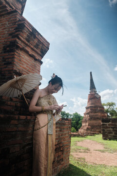 Cheerful Asian teenage girl tourist in traditional Thai outfit, social media sharing with mobile phone at old brick ancient ruins, holiday travel in Ayutthaya historic park,  travel destination.