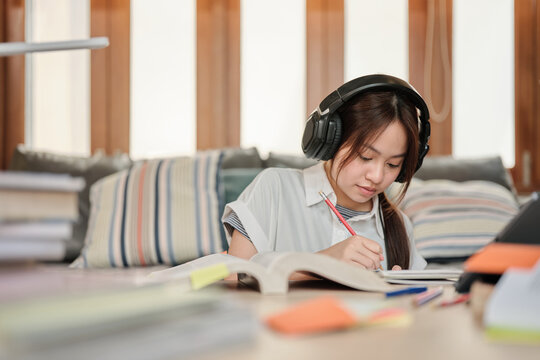 Asian teenage girl in headphones concentrates on learning at home, reading textbooks, note writing, self-tutorial exams, preparation for back to school, Homeschooling, university education lifestyle.