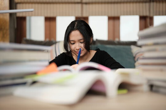 One Asian teenage girl concentrates on pile of books, focuses on learning at home with textbooks, cheerful reading and writing, self-study for school, Homeschooling, university education lifestyle.