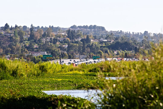 A green Santa Ana River corridor runs beside freeway traffic and hillside neighborhoods under clear summer skies in Yorba Linda.