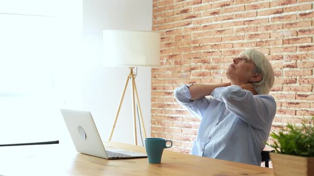 Exhausted senior business woman sitting at her desk in a modern office, experiencing severe neck pain and stiffness due to poor posture while working long hours on her laptop computer