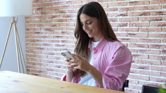A young woman happily engages with her mobile phone, browsing social media and smiling. She enjoys digital communication in the relaxed environment of her modern home.