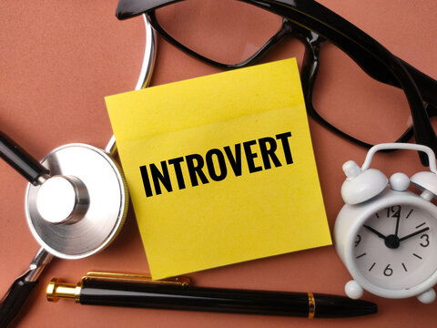 A yellow note labeled INTROVERT rests near a stethoscope, glasses, clock, and pen on a brown background