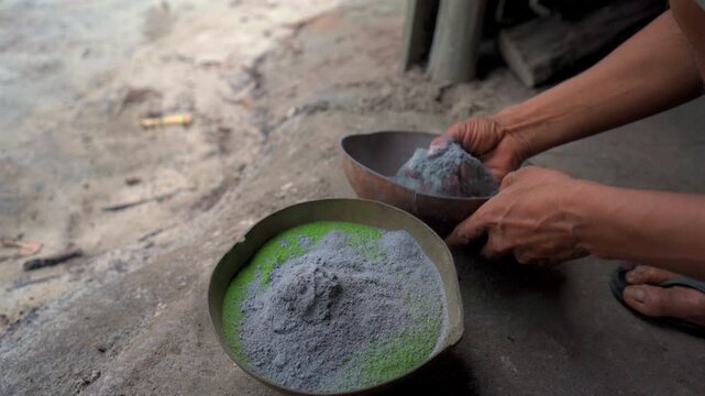 Static close up shot of indigenous hands mixing green mambe powder with grey ash in a gourd bowl on dirt floor inside an Amazonian maloca in Colombia.