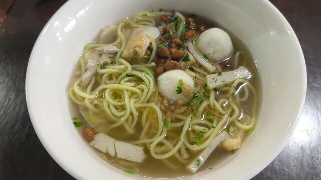 Asian noodle soup served hot in a white porcelain bowl. Yellow noodles with fish balls, fish cakes, and mixed vegetables in a clear light broth. Fresh and appetizing meal presentation.