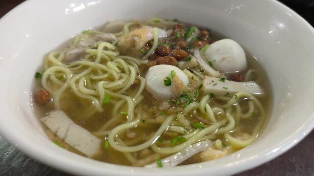 Asian noodle soup served hot in a white porcelain bowl. Yellow noodles with fish balls, fish cakes, and mixed vegetables in a clear light broth. Fresh and appetizing meal presentation.