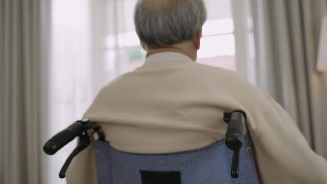 Unhappy senior Asian man is sitting alone in wheelchair and looking outside window due to depression and loneliness in his room at home. Sad elderly old paralytic male patient is disabled and lonely.