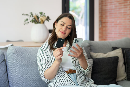 A smiling woman sits on a sofa at home, holding a smartphone and a credit card as she completes an online purchase. Casual, modern lifestyle scene conveying convenience and digital payment.