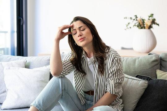 Young woman feeling intense headache or migraine, sitting on a comfortable sofa in a bright modern living room, experiencing stress and exhaustion from the discomfort