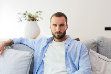 Fototapeta premium Young man sitting comfortably on a gray sofa in a bright, minimalist living room, looking directly at the camera with a calm expression, embodying a relaxed lifestyle