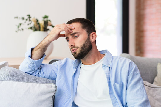A young man sits on a living room couch holding his forehead, appearing stressed and fatigued. Natural light and a calm interior contrast with his worried, pensive expression and signs of