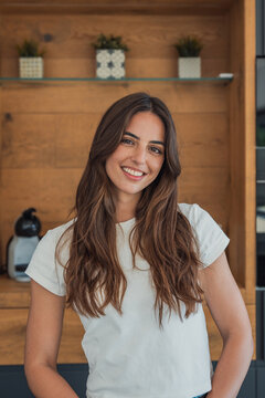 Smiling young woman standing in a modern wood-accented kitchen, wearing a white t-shirt. Natural, relaxed lifestyle portrait conveying warmth, confidence and everyday domestic comfort with a