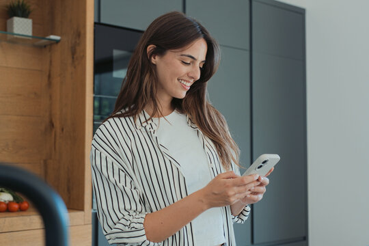 A smiling young woman stands in a modern kitchen, casually dressed and engaged with her smartphone. Warm, everyday lifestyle scene conveying connection, relaxation, and digital communication at home.