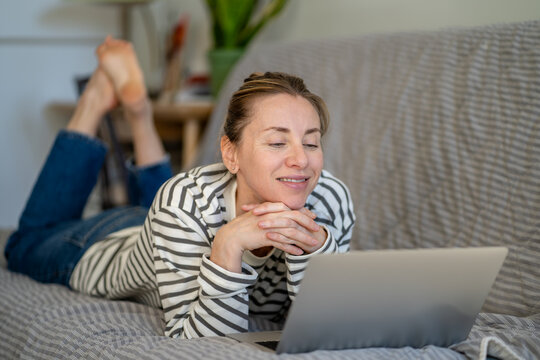 Happy relaxed woman lying on sofa at home resting and watching media content online on laptop, cheerful lazy female on sofa enjoying procrastination and free time on weekend, browsing on computer.