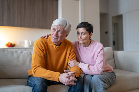 Compassionate wife providing silent support to distressed upset husband sitting on couch together, caring empathetic woman embracing and holding partner hand, senior couple struggle, emotional comfort