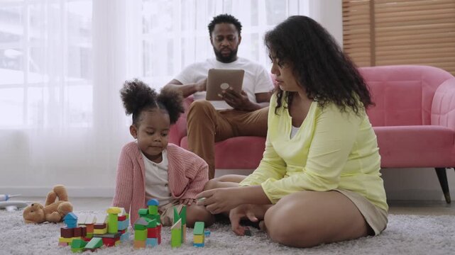 Family day concept. Happy African family is depicted with a mother and a cute little daughter playing with wooden blocks on the floor while the father sits on the sofa in the living room using tablet.